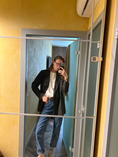 A young person with glasses, a white t-shirt, a dark blazer, and blue jeans is reflected in a set of mirrors in a room with textured yellow walls. There is a white air conditioning unit mounted high on the left wall. The room visible in the mirrors has a dark gray floor. The location is in Перепечино, Russia.