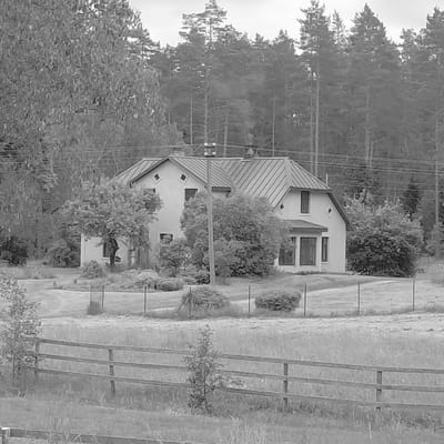 The black and white photograph depicts a rural scene in Garkalne, Latvia, featuring a house and its surrounding landscape. The house, a two-story structure with a sloped metal roof, is light-colored and appears to have diamond-shaped window accents. To the left of the house, a mature tree with dense foliage partially obscures the building. The foreground is characterized by a wooden fence with three horizontal rails. Beyond the fence, a grassy field with scattered vegetation stretches towards the house. In the background, a dense forest of tall coniferous trees forms a dark backdrop against a slightly overcast sky. Power lines are visible running horizontally across the upper portion of the image, strung between poles. The overall impression is one of a quiet, wooded residential area.