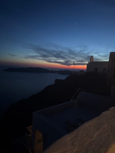 An outdoor, elevated shot captures a twilight scene in Thira Municipal Unit, Greece, likely during sunset or dawn. The sky transitions from a deep navy blue at the top to hues of orange and red near the horizon, with wispy clouds streaked across. In the foreground, the edge of a white-washed structure is visible, with part of a balcony or terrace and some lounge chairs suggesting a resort or residential area. Further out, silhouetted against the colorful sky, are the dark shapes of islands and cliffs overlooking a body of water. A white building with a steeple, resembling a church, stands on a cliff's edge, with a flag pole in front of it. Distant lights dot the coastline of the islands, indicating a settlement. Two small, bright points of light are visible in the upper right quadrant of the sky, possibly stars or distant lights. The overall atmosphere is serene and picturesque, characteristic of the Cycladic islands.