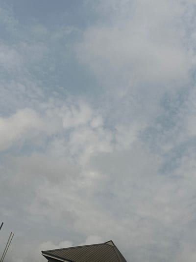 The image displays a sky predominantly filled with scattered white and light grey clouds against a pale blue background, occupying approximately the upper 80% of the frame. In the lower portion, the angular peak and slope of a dark brown or grey roof structure, featuring visible corrugations or ridges, are present. Two slender, vertical, pole-like objects are partially discernible along the extreme left edge, extending from below the image boundary. No individuals, specific actions, or interactions are depicted. The scene is located in Igwuruta, Nigeria.