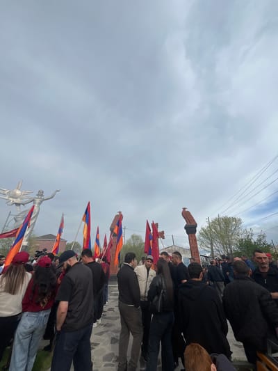 The image shows a crowd of people gathered in what appears to be a public space in the city of Akhuryan, Armenia. Several individuals are holding Armenian flags, and the scene features prominent statues. In the foreground, there is a statue of a woman standing in front of a sun. The monument to the martyrs is in the background, surrounded by Khachkars. People are congregated near these monuments. The sky is overcast.