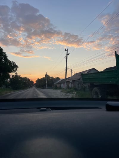 The image captures a street scene at dusk in Onițcani, Moldova. A dark, asphalt road stretches into the distance, lined on both sides by modest houses and trees. The sky above is a gradient of soft blue and orange hues, illuminated by the setting sun, with scattered, light pink clouds. Utility poles and power lines are visible throughout the scene. On the right side of the road, a green truck is parked, partially obscuring a row of houses. The foreground of the image shows the dark interior of a vehicle's dashboard. The overall atmosphere is tranquil and rural.