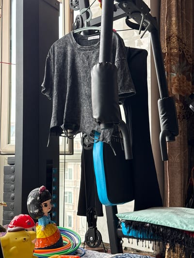 The image shows a cluttered indoor space with a home gym apparatus and various decorative items. A grey, acid-washed t-shirt hangs from a hanger on the gym equipment. To the left, a doll with black hair, a red bow, and a colorful dress sits on a pile of colorful hula hoops. A yellow, dog-like character with a red hat is also visible. The gym equipment has padded armrests and a black and blue padded seat. In the background, a window reveals buildings and a floral curtain. The scene is consistent with a domestic setting, possibly a home gym. The image is taken in Manzhouli, China.