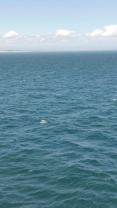 The image depicts a wide expanse of the ocean under a partly cloudy sky. The water is a deep blue-green and shows moderate choppiness with small waves. In the distance, a hazy coastline with a light-colored horizon is visible. Scattered across the water's surface are small patches of white foam or spray. The sky is a pale blue with fluffy white clouds near the horizon. The scene is devoid of any human presence or identifiable structures, suggesting an open water environment possibly off the coast of the United Kingdom, though no specific city is identifiable.