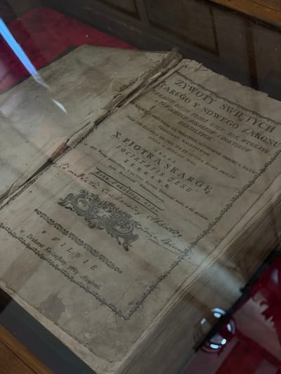 The image shows an open, antique book with yellowed pages and visible wear. The title page, written in an old script, is the main focus. The text appears to be in Polish, with phrases like "ŻYWOTY ŚWIĘTYCH" and "SKARGE JESTU". There is a decorative border around the text. Below the title, there are inscriptions and what appear to be handwritten signatures. The book is placed on a surface, possibly a display case, with reflections on the glass. The environment suggests an indoor setting, possibly a library or museum. The specific location in Літвенскі сельскі Савет, Belarus cannot be determined from the image.