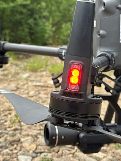 The image shows a close-up of a black drone with a red indicator light. The drone is on the ground, which is covered in small stones and patches of grass. In the background, there are blurred green trees. The drone appears to be a professional-grade model, possibly used for aerial photography or surveillance. The red light is on, suggesting the drone is active or preparing for flight. The focus is on the drone's arm and propeller. There are no people or animals visible in the image. The environment appears to be outdoors, possibly in a natural or semi-urban setting. The lighting suggests it is daytime. The location in Murtygit, Russia is not evident from the visual information in the image.