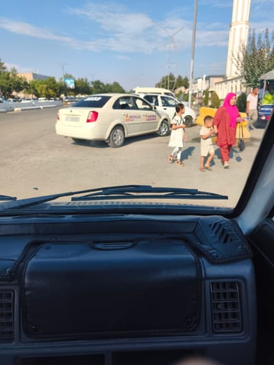 The image shows a street scene in the city of Buz, Uzbekistan, taken from inside a vehicle. In the foreground, the interior of the vehicle is visible, including the dashboard and windshield wipers. Outside the vehicle, a white taxi with "BONUS TAXI 2233" written on it is parked on the street. A woman in a pink hijab and a red outfit is walking with two children. The street is paved, and there are trees and buildings in the background. The sky is clear and blue.