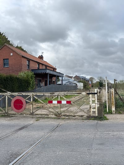 The image depicts a static outdoor scene, likely a rural railway crossing or a disused railway line in the Breckland District, United Kingdom, under an overcast sky. In the foreground, a weathered metal wire-mesh gate stretches across the frame, featuring a red and white striped barrier arm and a red circular sign with a white horizontal bar, resembling a "no entry" or "road closed" sign. Faint, illegible text is visible on the white bar. Two railway tracks are embedded within the paved ground, running from the bottom right towards the gate.
To the left of the gate, a prominent brick house with a tiled roof, a visible chimney, and a television aerial stands, partially enveloped by green hedges and bushes. Behind this, other residential buildings and structures are scattered along the railway line, interspersed with trees and additional fencing. The sky above is entirely covered by dense, grey clouds, indicating an overcast daytime. There are no people visible, and no specific activities are taking place, contributing to a quiet, somewhat still atmosphere. The aged appearance of the gate and surrounding infrastructure suggests a long-established or potentially disused line.