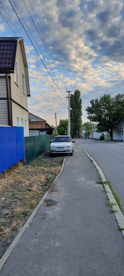 A light-colored sedan is parked on the side of a paved road in Shakhty City District, Russia. The car is positioned on the shoulder of the road, next to a sidewalk. A blue and green metal fence runs along the property line behind the car. To the left of the fence is a two-story house with beige siding and a dark brown roof. In the background, power lines crisscross the sky above a residential street lined with trees and other houses. The sky is partly cloudy with scattered white and gray clouds against a blue background. The scene appears to be a quiet, daytime residential setting.