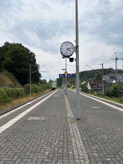 The image depicts a train station platform in Velbert, Germany. A green and white train is visible on the tracks, approaching the platform. A large clock with the "DB" logo is mounted on a pole in the foreground, indicating the time. Platform numbers "1" and "2" are displayed on blue signs. The platform itself is paved with grey bricks. To the left of the tracks, a grassy embankment is visible, covered in dense green trees. To the right of the platform, residential buildings with pitched roofs are nestled among trees. A construction crane is also visible in the background on the right. The sky is overcast with grey clouds.