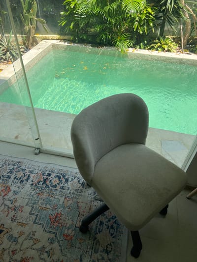The image presents a high-angle view of an interior space in Buduk, Indonesia, bordering an outdoor pool area. The pool's turquoise water contains some floating leaves. Lush tropical vegetation is visible beyond the pool. A beige office chair with black legs and a five-star base sits on a patterned rug with blue, orange, and grey tones. A glass barrier separates the interior space from the pool. The floor is tiled with light-colored stone. The lighting suggests daylight conditions.