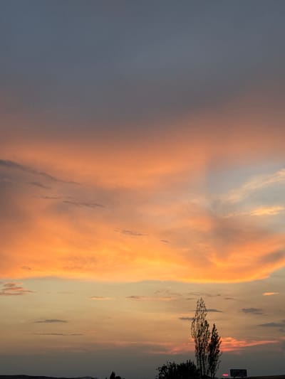 The image depicts a sunset over the city of Baş-Bulak, Kyrgyzstan. The sky is filled with dramatic clouds in shades of orange, pink, and grey. A silhouetted tree stands prominently in the foreground, with a signpost further to the right. The signpost displays Cyrillic text, which is partially visible. The overall scene is serene and captures the natural beauty of the landscape at dusk.
