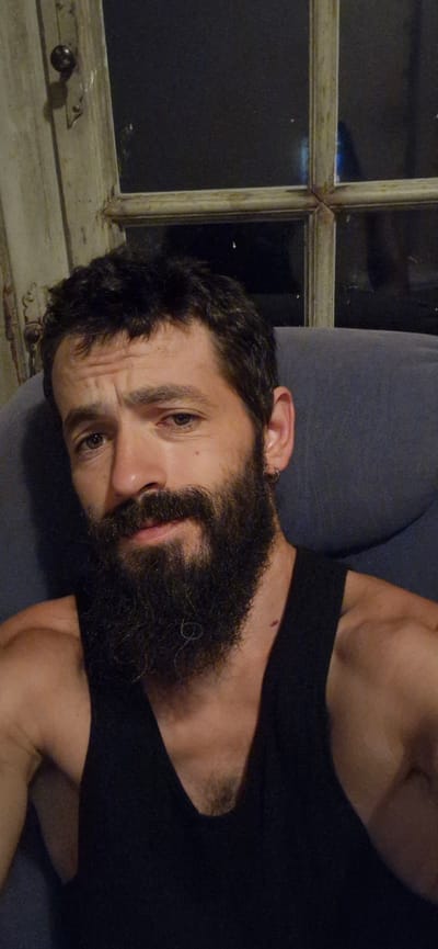 A man with dark, curly hair and a full beard is sitting in a dark room, possibly a living room. He is wearing a black tank top. In the background, there is a window with a wooden frame and glass panes. The window appears to be old and somewhat worn. The lighting in the room is dim, with the primary light source seemingly coming from outside the frame. The man is looking directly at the camera with a neutral expression. There are no visible signs of the location being in Wattenwil, Switzerland, nor any specific urban details. The scene is intimate and personal, focusing on the man's portrait.