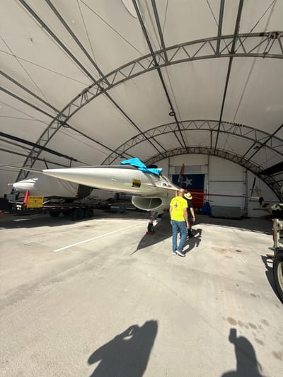 An aircraft sits within a large, semi-circular hangar with a webbed ceiling structure. A man stands near the aircraft's engine intake, wearing a yellow shirt, jeans, and a straw hat. The aircraft has a white nose cone, and a blue cloth is draped over part of it. The Texas state flag hangs in the background. A shadow of a person holding a camera is visible on the right side of the photo.
