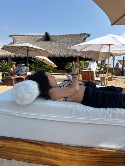 A woman with curly hair rests on a daybed with a white cushion, wearing a bikini top and black pants, appearing to look at her phone. In the background, a thatched roof structure and white umbrellas create a beachy ambiance. Other people and vegetation add to the outdoor scene under a clear blue sky.