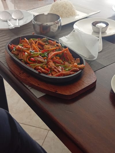 A dining table is depicted at an unidentified establishment in Murang'a, Kenya. An oval black cast iron sizzler plate, resting on a wooden trivet, occupies the foreground, filled with a stir-fried dish composed of julienned carrots, green and red bell peppers, red onions, and sliced meat or chicken pieces. To the right, a rectangular white plate holds a mound of white rice. Adjacent to the rice, a smaller white plate presents a small white bowl containing a dark sauce with a spoon. A crumpled white paper napkin and a cylindrical silver ashtray are positioned near the center of the table. On the far left, two white round plates with two large serving spoons are partially visible. The table itself is dark brown wood. The floor visible beneath the table is tiled with light-colored square tiles. A person's leg, clad in dark trousers, is partially visible in the lower-left corner of the frame.