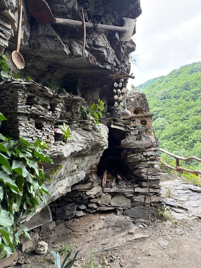 The image depicts a rustic dwelling or shelter integrated into a natural rock overhang in a mountainous region of Golyamo Belovo, Bulgaria. The primary geological feature is a large, grey, layered rock formation that forms a natural cave or deep recess.
Below the rock overhang, various structures have been built from stacked, flat grey stones, creating shelves, niches, and a chimney-like formation. A horizontal wooden beam is secured under the overhang, from which several items are suspended: a rustic wooden spoon, a rusted metal shovel blade, and a curved, rusted metal tool or hook. Also hanging from a chain attached to the beam is a decorative mobile composed of numerous small, white, shell-like objects or pebbles strung from a piece of driftwood.
Within the stacked stone structures, a dark ceramic pot and a white, gourd-shaped object are placed in niches, along with a small blue object. At the base of the stone structure, a fireplace area contains several logs. Leaning against this structure are two long, dark metal tools, possibly fireplace pokers or rakes, and a circular metal grill leans against a rustic wooden railing to the right.
The immediate ground is composed of dirt and smaller rocks, with patches of green vegetation including ivy clinging to the rock face on the left, succulents, and an agave-like plant on the ground. A small, light-colored rock carved or shaped like a face is visible on the ground to the left. In the background, lush green deciduous trees cover rolling hills under an overcast sky. No individuals or specific actions are depicted.