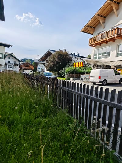 This image depicts a street scene in Kitzbühel, Austria, on a sunny day. The foreground is dominated by a weathered wooden fence and tall, green grass with some dandelion seed heads. Beyond the fence, a Billa supermarket is visible with its distinctive yellow signage. A white van is parked in front of the supermarket, and a few shopping carts are nearby.
The background shows several buildings, characteristic of Austrian architecture, with steeply pitched roofs. Some of these buildings have balconies with wooden railings. In the distance, rolling hills and mountains are visible under a clear blue sky with a few fluffy clouds. The overall atmosphere is peaceful and summery.
The time of day appears to be late morning or early afternoon, given the bright sunlight casting shadows. The weather is fair and warm. Visible text includes "BILLA," "WEINHERZ," and "Haus Schwager" on building signs. The scene conveys a sense of everyday life in a charming Austrian town.