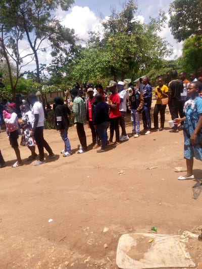 The image captures an outdoor scene in Mwingi, Kenya, featuring a group of approximately twenty to thirty people, mostly young adults, forming a loose queue on a dirt road. The individuals are dressed in casual attire, including t-shirts, shirts, jeans, and dresses, with some wearing caps and various types of footwear like sandals and sneakers. One woman on the left is seen holding an infant wrapped in a pink blanket, while another woman on the far right holds a white cup.
The people appear to be waiting in line, though the purpose of the queue is not evident. The overall mood seems calm and patient. The setting is characterized by a natural, slightly unkempt environment with numerous trees, some with dense green foliage and others with visible branches, suggesting a warm climate. In the background, partially obscured by trees, are small, simple buildings, possibly residences or community structures.
The ground is predominantly reddish-brown dirt, with scattered small pieces of litter, including a green plastic bottle and what appears to be a flattened bag or tarp in the foreground. The weather is bright and sunny, indicative of daytime, with a clear blue sky interspersed with white clouds. No discernible text is visible on any objects or clothing within the image.