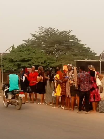 A group of young women is standing on the side of a paved road. Some are wearing dresses, and others are in skirts and blouses. A man on a motorcycle is driving past. Behind the women, a large tree spans the skyline, and other buildings and trees are in the background. The scene is bathed in soft, muted light, suggesting early morning or late afternoon.
