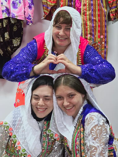 In Ribnovo, Bulgaria, three women pose for a photograph, each adorned in traditional Bulgarian attire. The woman at the top, her face beaming with a smile, forms a heart shape with her hands. She wears a white headscarf, a vibrant pink vest layered over a sparkling blue sequined top, and decorative gold trim. Beneath her, two women are positioned, also wearing white headscarves. The woman on the left, with her eyes closed and a joyful expression, is dressed in a heavily embellished garment featuring sequins and colorful floral embroidery. The woman on the right, with her eyes open and a wide smile, wears a similar richly decorated outfit with a blue satin top and lace sleeves. Behind them, colorful, patterned fabrics hang, suggesting a backdrop of traditional clothing or textiles.