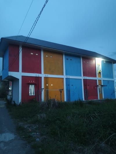 This is a medium-shot, eye-level, outdoor photograph taken at dusk in Panteraya, Indonesia. The main subject is a brightly colored, multi-story building with sections painted red, yellow, and blue, divided by white structural elements. The building appears to be an apartment complex or dormitory. Several small, square windows with ventilation patterns are visible on the upper floors. One window on the lower left is illuminated from within. On the right side of the building, a person wearing a yellow shirt is visible in an upper-story opening, appearing to be working or looking out. The sky is a deep blue, indicating the time of day is twilight, and there are no visible weather conditions. The foreground consists of overgrown grass and a dirt path leading to the left of the frame. The overall mood is quiet and somewhat subdued due to the low light. There is no visible text in the image.