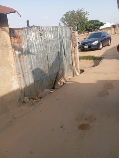 The image depicts a street scene in Gwagwa, Nigeria. On the left, a roughly constructed wall of corrugated metal is visible, showing signs of rust and damage. Adjacent to it is a light-colored, stucco-like wall. A portion of a reddish-brown, corrugated metal roof juts out from the top left of the frame. The street itself is unpaved, composed of dirt and sand, with some scattered rocks and debris. In the background, a dark-colored sedan, possibly a Honda Accord based on the license plate which reads "ABC 365JZ", is parked on the right side of the road. Beyond the car, there are some low-lying buildings with concrete walls and a few trees with green foliage under a clear, bright sky. The overall impression is of a modest, semi-urban or rural environment.