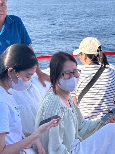 In Dong Son, Vietnam, on a boat amidst a body of water, a group of individuals are engaged in various activities. A man in a blue collared shirt stands in the background. In the foreground, two young women are seated, both wearing face masks. The woman on the left, with dark hair pulled back, is wearing a white top and looking down. The woman in the center, also with dark hair, is wearing glasses and a light green top, and is holding a smartphone. Another woman, in the background, is wearing a white baseball cap and a striped shirt, also holding a smartphone. The boat appears to have a red railing. The water is blue and appears to be a sea or large lake.