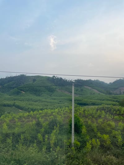 The image shows a landscape in Thôn 18, Vietnam, with a hillside covered in dense green vegetation. A utility pole stands prominently in the foreground, with power lines extending across the scene. The sky is overcast with bright white sunlight shining through. The terrain appears hilly and forested. No people are visible.