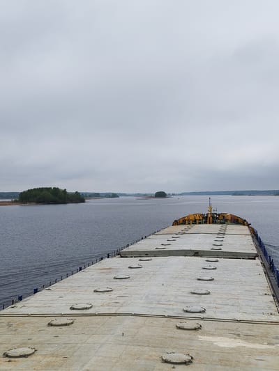The image shows the deck of a ship moving along a body of water under a cloudy sky. The ship's deck is a flat, gray surface with circular hatches arranged in rows. In the distance, there are islands with green vegetation and hints of land on the horizon, suggesting a wide river or lake. The location is Staroye Melkovo, Russia.