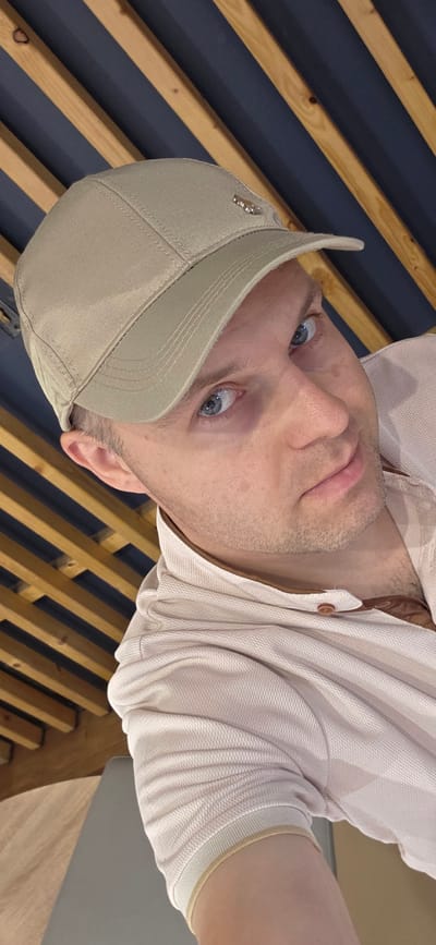 The image is a close-up, high-angle selfie of a man wearing a light-colored baseball cap and a light-colored polo shirt. He has blue eyes and light skin with some stubble. The background consists of wooden slats with dark blue in between, arranged diagonally. The man's right arm and shoulder are visible, and he is looking directly at the camera. The overall lighting suggests an indoor setting. There is no visible indication of the specific location within Collado Villalba, Spain.