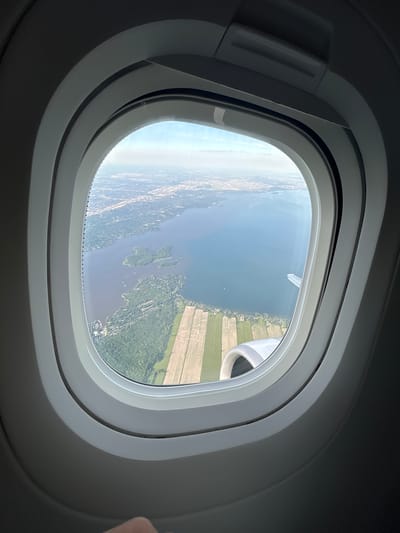 An aerial view from an airplane window shows a sprawling urban landscape bordering a large body of water. The foreground is dominated by the rounded, gray interior of the airplane cabin and window frame. The window looks out onto a scenic vista featuring a wide expanse of blue water, dotted with small boats. Along the shoreline, there are areas of dense green forest and agricultural fields with a distinctive pattern of alternating light and dark strips, suggesting plowed or planted land. In the distance, a city with buildings and infrastructure stretches towards the horizon under a hazy sky. The wing of the airplane is partially visible, extending out over the water. The image captures a typical scene experienced during air travel, offering a perspective of the earth from above.