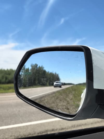 This image shows a view through a car's side mirror. The mirror reflects a clear blue sky with wispy white clouds. Below the sky, the reflection shows a rural highway with a white line marking the lane. A light-colored car is visible driving in the distance on the highway. To the left of the highway in the reflection, there is a dense line of green trees. The mirror itself appears to be somewhat dirty with smudges and water spots. The exterior of the car the mirror is attached to is visible on the right side of the frame, appearing white and slightly angled. The overall scene suggests a bright, sunny day on a road trip through a rural area. The location is identified as Prikhodnyy Aul District, Kazakhstan.