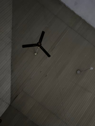 The image depicts a ceiling view within an interior space, likely in Abeokuta, Nigeria. The ceiling has a sloped, angular structure, constructed from ribbed panels, with a light gray color. A ceiling fan with three black blades is situated on the left side. A single bare light bulb, affixed to a small circular fixture, is present on the upper right. The overall lighting is dim.