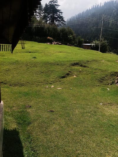 The image depicts a grassy slope in Khakhnal, India. In the foreground, the edge of a wooden structure, possibly a porch or balcony, is visible on the left. The slope is covered in short, green grass, with some uneven patches and scattered small stones. In the midground, a small, rustic wooden building with a tin roof is situated amongst trees, with power lines running overhead and a utility pole nearby. Behind this, a dense forest of coniferous trees covers a mountain slope, receding into a misty, overcast sky. In the distance, power lines extend across the landscape. There are no people or animals visible in the scene.