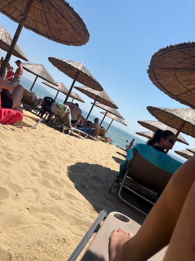 The image depicts a sunlit beach scene in Logkari, Greece, observed from the low perspective of someone reclining on a sunbed. The foreground shows the bare feet and lower legs of the viewer, propped on a light-colored lounge chair. The sand is golden-brown, displaying ripples and small indentations.
Numerous straw beach umbrellas (paliotas) are scattered across the mid-ground and background, providing shade. Underneath these umbrellas, multiple individuals are relaxing on beige-colored sunbeds with metal frames.
In the right mid-ground, a female adult is seen in profile, wearing dark sunglasses and a dark top, with her dark hair tied back. She is reclining on a sunbed draped with a bright blue towel. Towards the left mid-ground, another individual's lower torso and legs, partially covered by a pink or red garment, are visible on a sunbed.
Further into the scene, a shirtless male adult wearing patterned swim shorts stands near an umbrella, seemingly looking down at something in his hands. Nearby, another sunbed holds a towel with cartoon characters. In the background, several people are visible in the calm, blue sea, some wading or swimming. Distant landmasses or mountains are faintly visible across the horizon of the water. The sky above is clear and bright blue, indicative of a sunny day, with strong sunlight casting distinct shadows from the umbrellas and people.