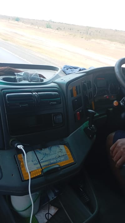 The image displays the interior of a vehicle, presumed to be a truck, viewed from the passenger side looking forward through the windshield. The vehicle is in motion within Malewa West ward, Kenya, on a two-lane paved road that traverses a dry, light-brown landscape characterized by sparse scrub vegetation and distant hills under a bright, hazy sky.
The vehicle's dark grey dashboard is prominent in the foreground. On the left, a cupholder contains a tuft of dark hair and clear plastic packaging. A blue microfiber cloth is draped over the dashboard on the right. The central console features air vents, a storage compartment, and a 12V power outlet. Below this, a rectangular panel covered with blue and yellow reflective material is present, over which a white USB charging cable, plugged into a cigarette lighter adapter, extends. Below the dashboard, a white bucket with a green handle rests on the floor on the left.
On the right side of the frame, a segment of the steering wheel is visible, along with a portion of the instrument cluster displaying gauges. A section of the dashboard is equipped with various buttons, switches, and a black-knobbed gear shifter. The driver's right hand and a segment of their right leg, clad in dark shorts, are visible on the extreme right, indicating active operation of the vehicle.