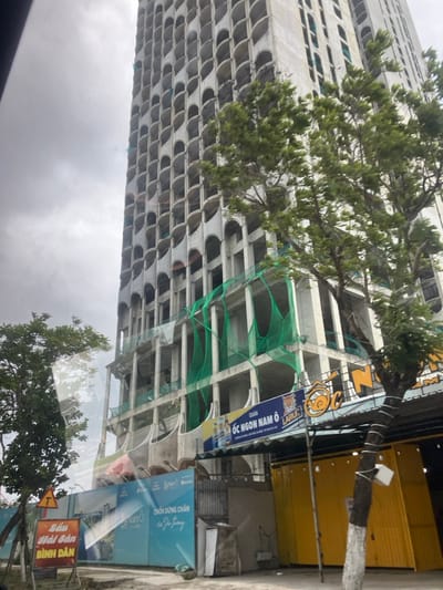 The image depicts a tall, multi-story concrete building under construction, viewed from a street-level perspective in Hải Vân Ward, Vietnam. The building's facade features a repeating pattern of numerous arched openings, suggesting balconies or window frames. Green safety netting is visible draped over several lower and mid-level sections of the unfinished concrete structure, particularly around exposed floor plates and columns.
In the foreground, a blue construction hoarding runs horizontally, displaying Vietnamese text including "Nam Ô" and "Chốn dừng chân của Gia Trung" alongside an architectural rendering. To the right of the hoarding, at street level, is a commercial establishment with a prominent blue sign bearing the text "QUÁN ỐC NGON NAM Ô" (Snail Restaurant Nam Ô) and a snail logo, along with "Larue" beer branding. Below this sign are several yellow, container-like structures and a covered outdoor dining area.
A tree with green foliage stands in the mid-ground to the right, partially obscuring the building and the commercial area. Another smaller tree is visible on the left foreground, near a yellow triangular road sign with a black 'T' and a red rectangular sign with Vietnamese text "CẦU HẢI VÂN ĐÌNH ĐÂN". The sky is overcast and grey. No individuals are visible in the scene, and no specific actions or interactions are depicted.