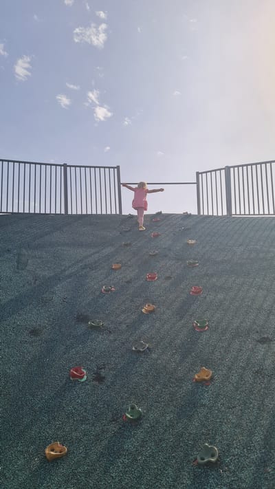 A child in a pink outfit balances on a climbing wall at a playground in Kiryat Gat, Israel. The climbing wall is grey with colorful climbing holds, and surrounded by a black metal fence. The sky above is blue with white clouds.