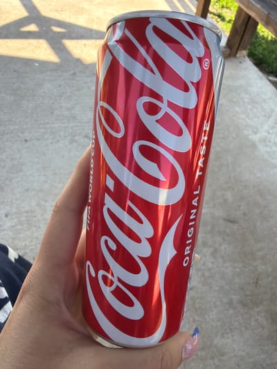 A person's hand holds a red Coca-Cola can with white lettering. The can has "FIFA WORLD CUP" and "ORIGINAL TASTE" printed on it. The background appears to be an outdoor concrete surface with shadows cast by unseen objects, possibly indicating a sunny day. There is some greenery visible in the upper right portion of the image. The location is not identifiable as Dinkata, Bulgaria.