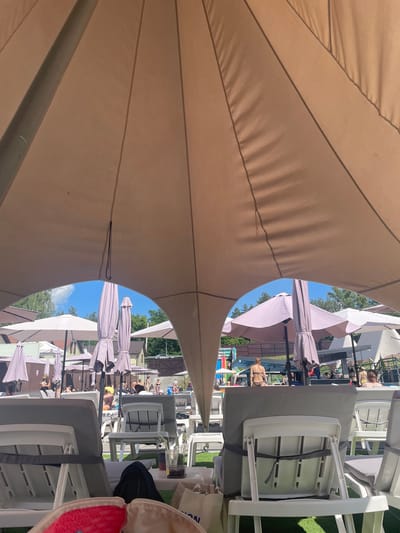 The image depicts an outdoor area with lounge chairs and umbrellas, suggesting a resort or recreational facility. The foreground features several white and grey lounge chairs covered by a large, beige, tent-like canopy, providing shade. In the background, under a clear blue sky with scattered clouds, more umbrellas and lounge chairs are visible. People are scattered throughout the area, some seated on chairs and others standing. Some individuals are dressed in swimwear, indicating a poolside or beach environment. Buildings, trees, and a playground structure are also present in the distance. The scene appears to be a sunny day, with artificial light sources not clearly visible. The location is identified as Zazymia, Ukraine, suggesting a summer scene at a place like the "Zazymia Resort and Spa".