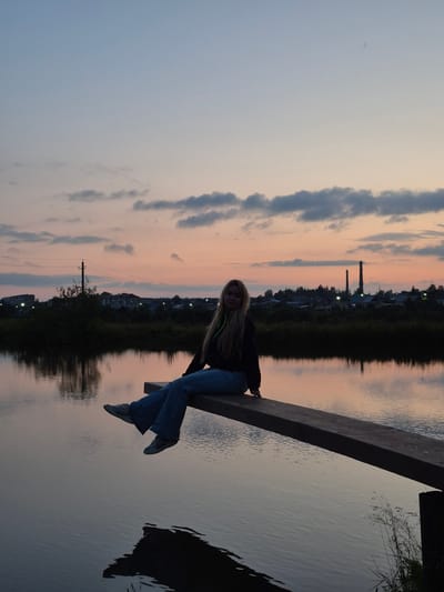 A young blonde woman with long hair is sitting on a wooden beam that extends over a body of water. She is wearing a dark jacket and blue jeans. The woman is leaning back slightly, with her legs dangling over the water. The water reflects the colors of the sunset. The sky is a gradient of pale blue to soft pink and orange, with scattered clouds. In the background, there is a dark line of trees and buildings, with some lights visible. Two industrial smokestacks are also visible in the distance, as well as power lines. The overall scene suggests it is dusk. The water appears calm. There is no visible text in the image.