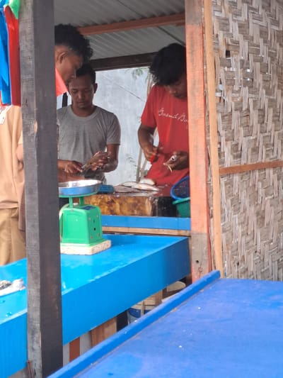 In Cottarum Tunong, Indonesia, three individuals are engaged in a fish preparation activity within a makeshift market stall. The stall has a corrugated metal roof and walls constructed from woven bamboo. In the foreground, a bright blue counter dominates the scene, with a green weighing scale positioned on it. A person in a grey t-shirt is seen holding a fish and a knife, seemingly in the process of cleaning or preparing it, with a metal bowl on the scale. Another person, wearing a red t-shirt with the inscription "Suma Bora," is also visible behind the counter, holding a knife and a fish, suggesting they are also involved in the fish preparation. A third individual, whose face is partially obscured, stands behind the person in the grey t-shirt. Several fish are laid out on a wooden surface in the background, and a green basket is also present. The overall environment suggests a bustling local marketplace where fish is being processed and sold.