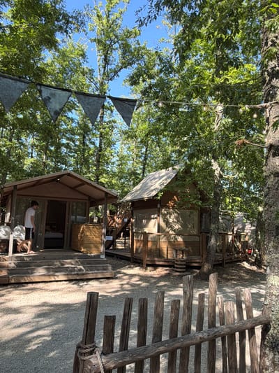 The image depicts a glamping site nestled within a wooded area, under a bright blue sky. In the foreground, a rustic wooden fence made of uneven posts and a horizontal beam, secured with rope, borders a gravel path. Beyond the fence, two wooden structures resembling cabins with canvas roofs are visible. The structure on the left has an open doorway revealing a bed inside and a person in a white t-shirt and shorts walking on the wooden porch. To the right of this structure, a wooden bin stands on the porch. The cabin on the right features a wooden deck with stairs leading up to it, and a railing surrounds the deck. String lights are suspended between trees, and triangular metallic flags hang above the cabins. The ground is covered with gravel and dappled with sunlight and shadows cast by the surrounding trees, whose lush green foliage dominates the background. The scene suggests a tranquil outdoor setting, likely a resort or campsite, possibly in the vicinity of Cavriglia, Italy, given the context.