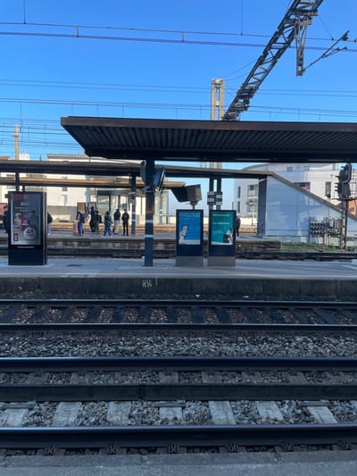 The image displays a daytime scene at a train station in Saint-Cyr-l'École, France, captured from the perspective of an adjacent platform or track level, looking across to an active passenger platform.

In the foreground, four parallel railway tracks are visible, consisting of metal rails, a mix of wooden and concrete sleepers, and light gray gravel ballast. A concrete platform edge, bearing the white painted alphanumeric designation "R16," separates the foreground tracks from the main platform.

On the main platform, approximately ten individuals, presumed to be waiting passengers, are standing. They are predominantly clustered under a dark, ribbed overhead canopy supported by several dark vertical pillars. One individual near the left of the group is positioned beside a light-colored rolling suitcase. Three prominent vertical digital display screens are present: one large advertising display on the far left featuring French text ("RELAIS," "4€ OFFERTS"), and two central screens exhibiting blue and green advertisements, one titled "SANS SMARTPHONE" and the other "AVEC SMARTPHONE." An extensive network of overhead electrical catenary lines and support gantries for the railway system is visible across the midground and background sky.

In the background, beyond the platform, additional railway lines are discernible. Multiple modern buildings with white facades and varied rooflines are visible, indicating urban or suburban development. To the far right, a sloped gray structure, possibly a station amenity or access point, and a vertical railway signal pole with multiple black signal lights are present. The upper portion of the image is dominated by a clear, bright blue sky. Minimal vegetation is observed along the track and platform edges.