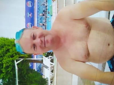 The image shows a man wearing blue swimming goggles, shirtless and with his head tilted back. He appears to be in a semi-tropical environment, with lush green foliage visible on the left side of the frame. Behind him, a swimming pool is visible, with several people in the water. A large blue banner with the Pepsi logo is displayed along the edge of the pool. The man's body shows signs of sun exposure, with areas of redness on his chest and arms. The overall impression is that of a vacation setting, likely at a resort or hotel with a swimming pool. There is no specific indication of the location being within the city of Birlik, Uzbekistan.