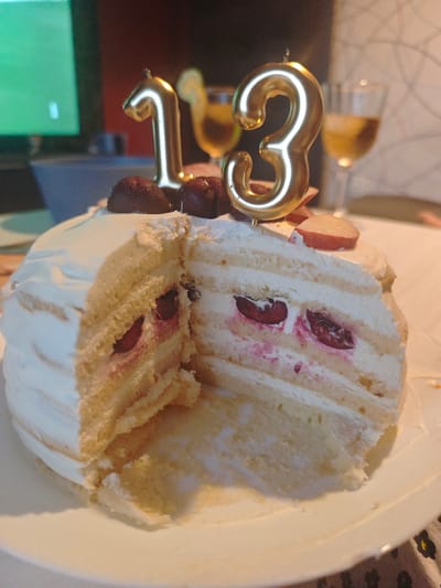 A close-up, eye-level shot shows a slice of a multi-layered white cake decorated with golden candles shaped like the numbers "1" and "3", indicating a birthday celebration for someone turning thirteen. The cake, placed on a white plate, features layers of sponge cake interspersed with white cream and visible whole cherries. The cream is swirled and covers the exterior of the cake. Some cherries and pink juice have seeped into the cake layers. Dark red, whole fruits, possibly cherries or chocolates, are arranged on top of the cake, along with thin slices of red fruit, possibly apples. In the background, a glass of amber liquid, likely wine or juice, and another out-of-focus glass are visible. A television screen displaying a green hue is also present to the left. The scene appears to be indoors, possibly in a residential setting in El Puerto de Santa María, Spain, given the context of previous interactions. The lighting is dim, with the cake and candles being the primary focus.