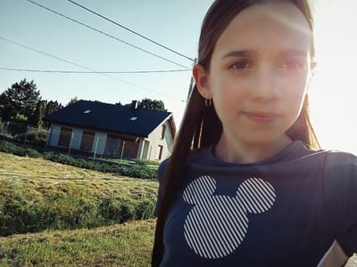 A young girl with long brown hair takes a selfie outdoors. She is wearing a dark blue t-shirt with a white striped Mickey Mouse design on the chest. The girl is positioned on the right side of the frame, with her face close to the camera. Her expression is neutral. A golden hoop earring is visible in her left ear.
In the background, a house with a dark roof and light-colored walls is visible. The house has several windows with brown shutters. A grassy yard with some vegetation and trees surrounds the house. The scene appears to be in a rural setting. Several power lines are strung across the sky.
The lighting suggests it is daytime, likely late afternoon or early evening, with the sun casting a warm glow and creating some lens flare on the right side of the girl's face. The sky is clear and light blue. The location is Dybów Kolonia, Poland. No text is visible in the image.
