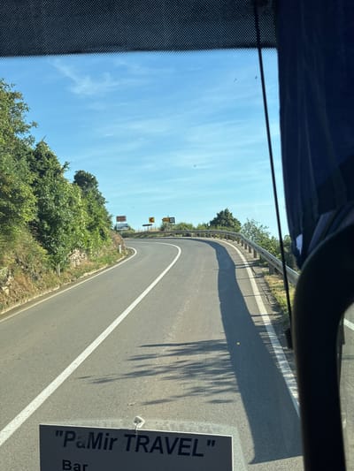The image captures a view from a vehicle, likely a bus, traveling on a winding road in the city of Blizikuće, Montenegro. The road curves to the left, bordered by a metal guardrail on the right and a steep, tree-covered embankment on the left. The sky above is a clear blue with a few wispy clouds. Road signs are visible in the distance, indicating curves in the road. The lower portion of the image shows a sign that reads "\"PaMir TRAVEL\" Bar". The overall scene depicts a scenic mountain road in a coastal region, suggesting a tourist or travel context.