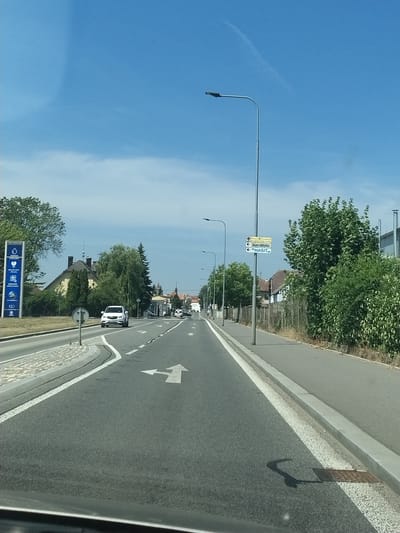 This is a ground-level, eye-level, wide shot of a paved road in Horažďovice, Czechia, on a bright, sunny day with a clear blue sky and scattered clouds. The road curves slightly to the right and recedes into the distance. On the left side of the road, there is a sidewalk, a grassy area, and trees, followed by a yellow building and a large blue sign with white text and icons, likely for a service station or gas station. A white car is driving on the road in the same direction as the viewer. Lane markings, including a left-turn arrow, are visible on the road surface. On the right side of the road, there is a sidewalk, a fence, and dense green foliage. Several streetlights are visible along the road. Further down the road, there are more buildings and trees. A directional sign with yellow and white text is mounted on a pole. The scene appears to be a suburban or semi-rural area.